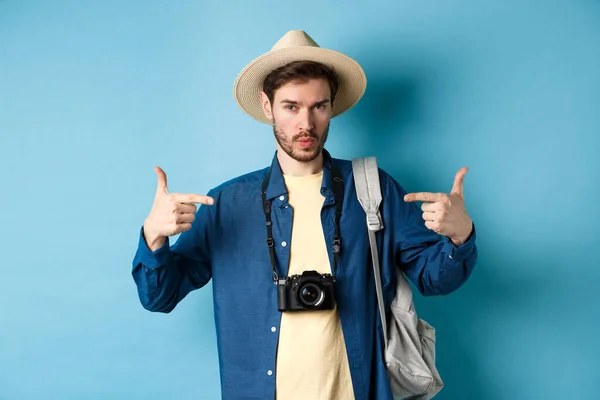 Jovem confiante e pronto apontando para si mesmo, auto-promoção ou show-off, indo de férias de verão, usando chapéu de palha, segurando mochila com câmera, fundo azul — Fotografia de Stock