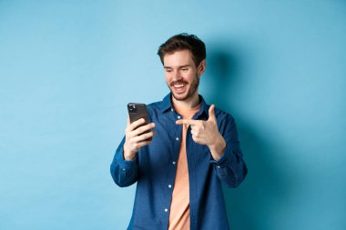 Mobil ekranı okuyan, telefonu işaret eden, online tanıtım ya da akıllı telefon uygulamasını kontrol eden, mavi arka planda duran yakışıklı bir adam resmi