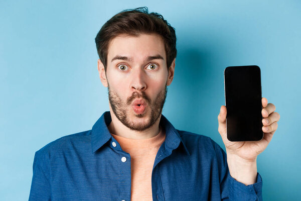 Close up portrait of impressed man say wow, showing empty mobile phone screen, standing on blue background