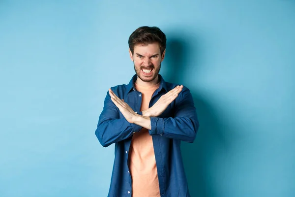 Злой молодой человек хмурясь и сжимая зубы возмущен, показывая крест жеста, чтобы остановить или запретить что-то, стоя на синем фоне — стоковое фото