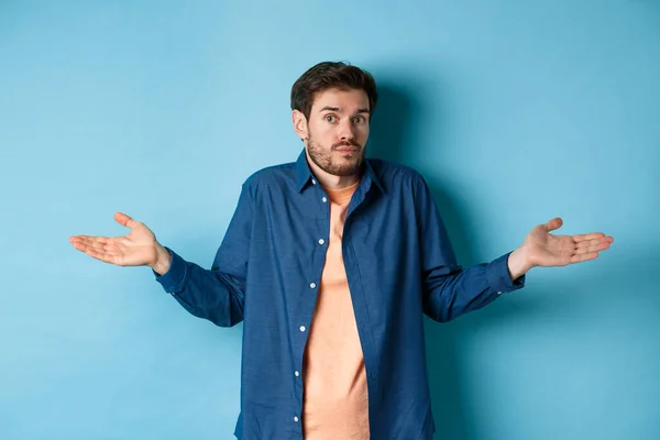 Verward jongeman met baard, schouders ophalen en op de camera kijken, niets weten, op een blauwe achtergrond staan — Stockfoto