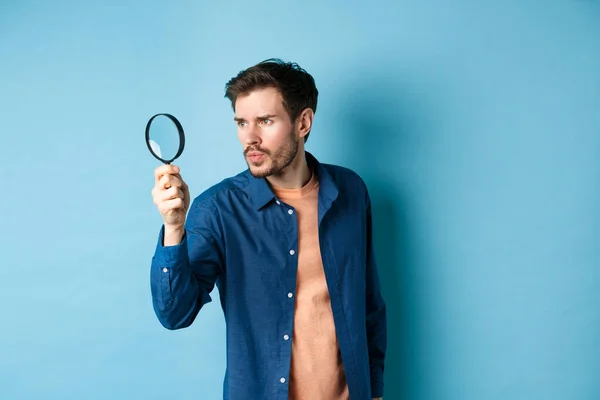 Serious man investigating or searching for something, look through magnifying glass like detective, standing on blue background — Stock Photo, Image