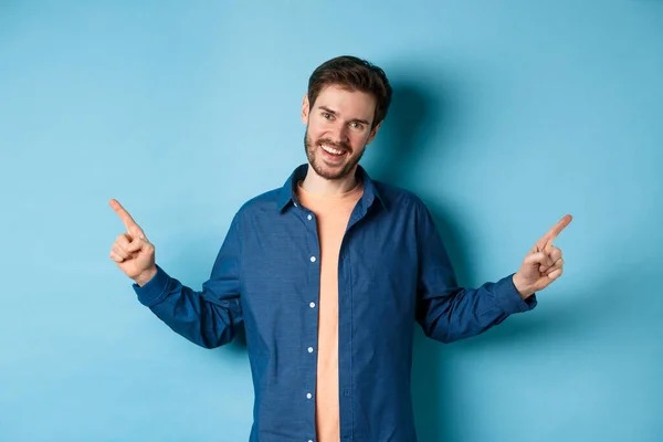 Handsome caucasian man with beard, smiling with white teeth and pointing fingers sideways, showing two choices, standing on blue background — Stock Photo, Image