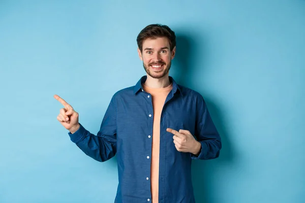 왼쪽 손가락을 로고로 가리키며, 광고를 보여 주고, 파란 배경 위에 서 있는 행복 한 미소짓는 남자 — 스톡 사진