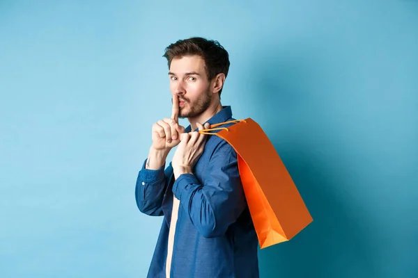 Image of modern guy holding shopping bag on shoulder, turn behind and shushing at camera, telling a secret, making surprise, standing on blue background — Stock Photo, Image