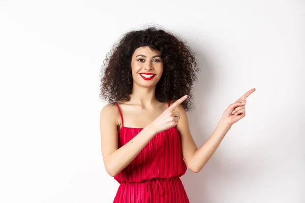 Vrolijk vrouwelijk model in modieuze rode jurk, glimlachend en wijzend vingers recht naar logo, staande over witte achtergrond — Stockfoto
