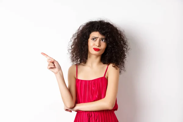 Mujer joven decepcionada y escéptica con el pelo rizado, vestido rojo, mueca y el dedo que apunta a la izquierda en el logotipo, de pie sobre el fondo blanco — Foto de Stock
