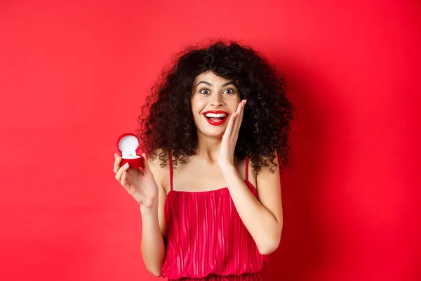 Beeld van verrast jonge vrouw met krullend kapsel, het dragen van rode jurk en lippenstift, tonen verlovingsring, gaan trouwen, staan op studio achtergrond — Stockfoto