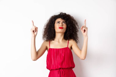 Kızgın ve huysuz bir kadın, kıvırcık saçlı, kırmızı elbiseli, kaşlarını çatmış, hayal kırıklığına uğramış, beyaz arka planda duruyor.