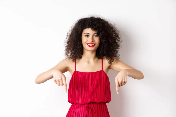 Attraente modello femminile in abito rosso e trucco, puntando il dito verso il basso e sorridente, mostrando pubblicità, in piedi su sfondo bianco — Foto Stock