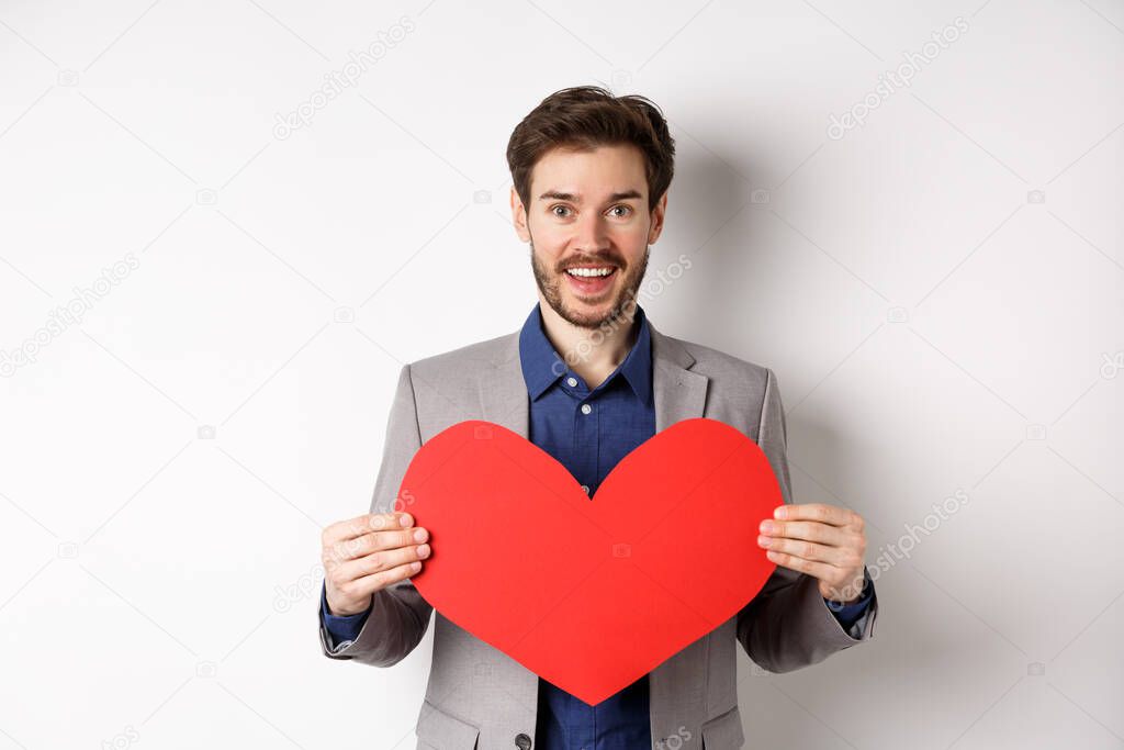 Happy man confessing in love, showing heart cutout and smiling at camera, standing in suit on romantic date with lover, white background