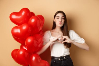 Sevimli kız arkadaş dudaklarını büzüyor ve romantik bir öpücük bekliyor, seni seviyorum demek için kalp jesti yapıyor, sevgiliye bak, sevgililer günü balonları ve bej arka plan