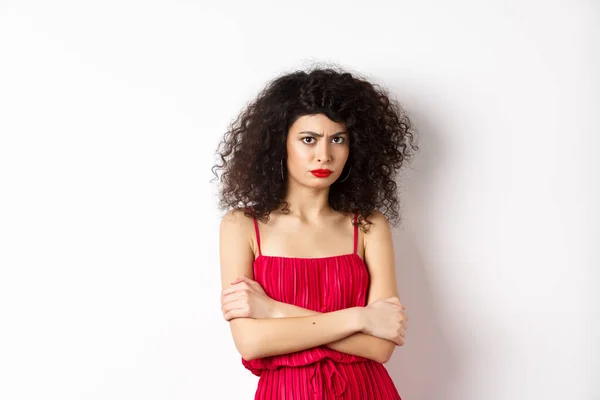 Sad and offended caucasian woman in red dress frowning, cross arms on chest and sulking, feeling mad at you, standing over white background — Stock fotografie