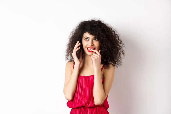 Глупая модная женщина в красном платье и губах, кусающая ногти дуя телефонный звонок, думая о мобильном разговоре, стоя на белом фоне — стоковое фото