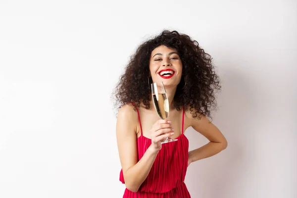 Donna allegra ed elegante con i capelli ricci scuri, indossa un abito da festa, celebra l'anniversario il giorno di San Valentino, beve champagne dal vetro e sorride alla macchina fotografica, sfondo bianco — Foto Stock