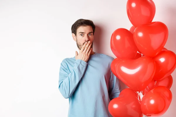 Retrato del novio hombre de pie cerca de los globos cardíacos de San Valentín y jadeando conmocionado, de pie sobre el fondo blanco en cuestión —  Fotos de Stock