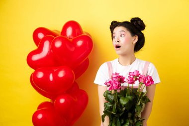 Sevgililer günün kutlu olsun. Şaşırmış Asyalı kız sol tarafa bakıyor, elinde sevimli pembe çiçekler, romantik bir buket alıyor, sarı arka planda kırmızı kalp balonlarının yanında duruyor.