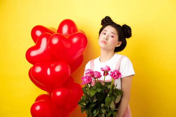 Счастливые Валентины. Грустная и мрачная азиатка, держащая букет роз и чувствующая себя расстроенной и одинокой в день романтических влюбленных, стоящая рядом с красными сердечными шарами, желтым фоном — стоковое фото