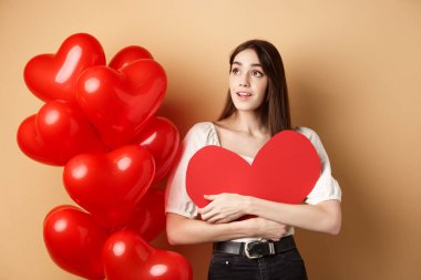 Romantik kız Sevgililer Günü 'ne sarılıyor, büyük kırmızı kalp kesiği, romantik balonların yanında duruyor ve logoya bakıyor, reklamlara bakıyor, bej arka planda hayaller kuruyor.