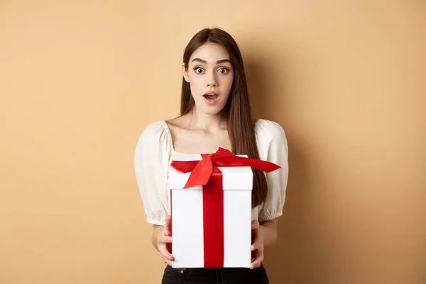 Amazed beautiful girl receiving gift from secret admirer on Valentines day, looking surprised at camera, holding box and standing on beige background