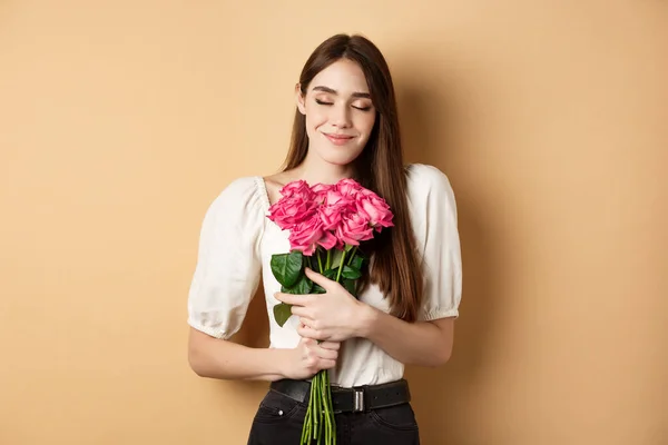 Día de San Valentín. Mujer blanca tierna y romántica ojos cerrados soñadora, abrazando ramo de rosas de admirador secreto o amante, de pie sobre fondo beige — Foto de Stock