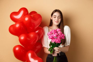 Gülleri koklayan romantik kız arkadaş Sevgililer Günü 'nde sevgililerinden çiçek alır kırmızı kalplerin yanında durur balonlar ve gülümser, bej arka plan
