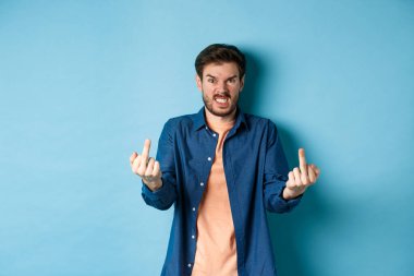 Angry rude guy showing middle finger and saying fuck you, swearing and staring furious at camera, standing on blue background