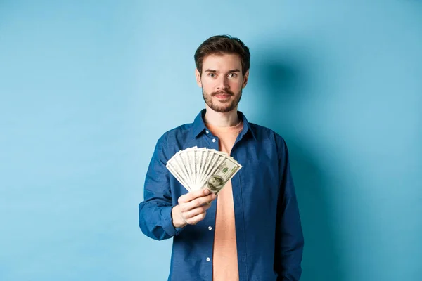 Feliz chico moderno sosteniendo el dinero y sonriendo, de pie sobre fondo azul — Foto de Stock