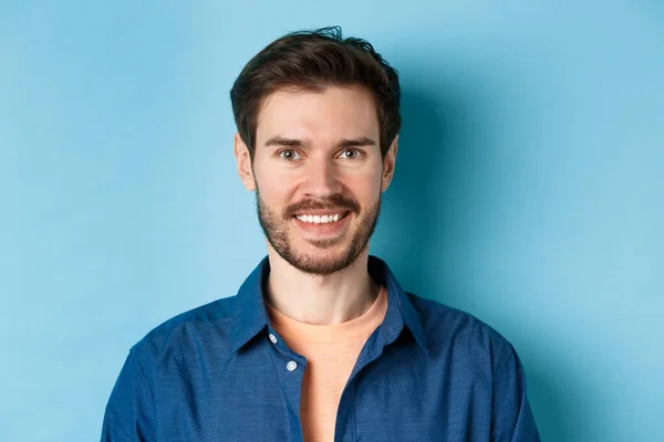 Nær lykkelig og sunn ung mann med skjegg smilende foran kamera, stående på blå bakgrunn – stockfoto