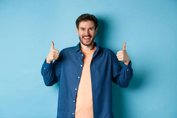 Vrolijke blanke man in casual shirt tonen duimen omhoog in goedkeuring, glimlachen en prijzen iets goeds, staan op blauwe achtergrond — Stockfoto