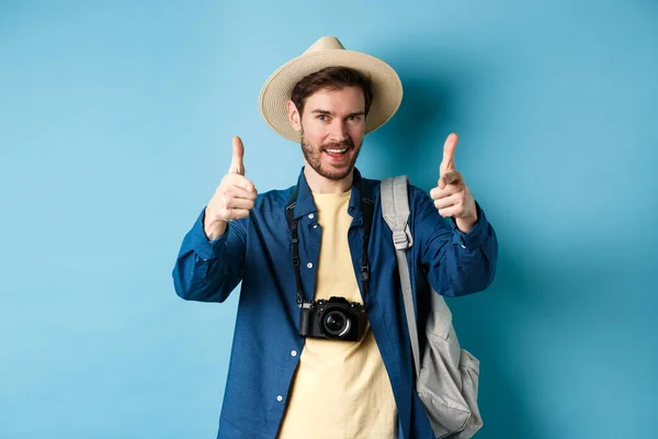 Turista bonito indo de férias de verão com câmera e mochila apontando os dedos para a câmera e convidando você, de pé sobre fundo azul — Fotografia de Stock