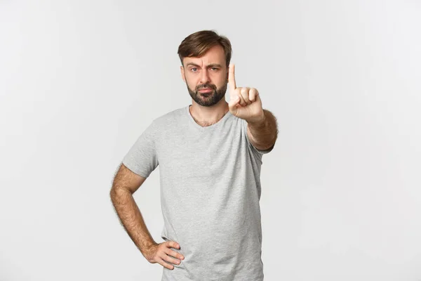 Portrait d'un homme adulte sérieux avec barbe, portant un t-shrit gris, secouant le doigt avec un visage déçu, grondant quelqu'un et interdisant l'action, debout sur un fond blanc — Photo