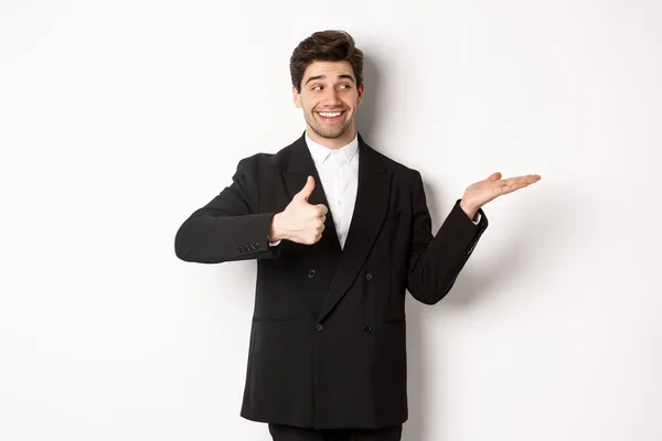 Snygg affärsman i svart kostym, visar tummen upp och hålla din produkt i handen över vit kopia utrymme, står mot vit bakgrund — Stockfoto