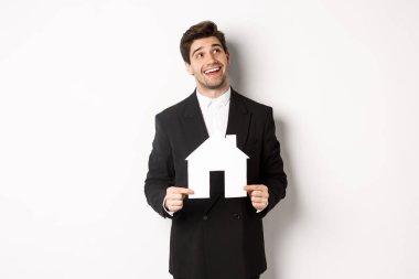 Evini arayan takım elbiseli yakışıklı bir adamın portresi. Elinde kağıt bir ev, sağ üst köşedeki hayallere bakıyor. Beyaz arka planda duruyor.