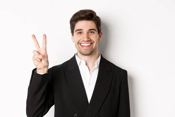 Närbild av stilig affärsman i svart kostym, leende förvånad, visar nummer två, stående över vit bakgrund — Stockfoto