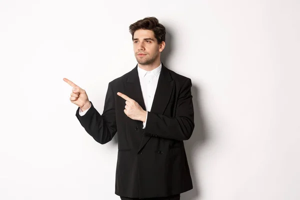 Porträtt av allvarlig stilig man i svart kostym, pekar och tittar vänster på promo banner, visar reklam på kopia utrymme, står över vit bakgrund — Stockfoto