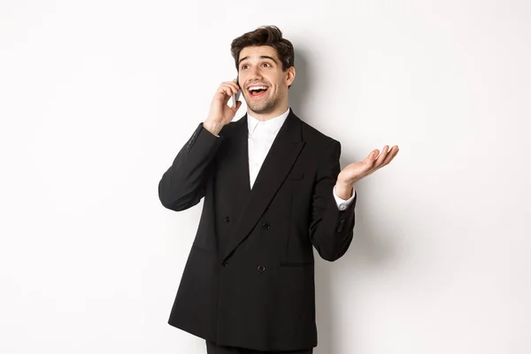 Portret van een blije, goed uitziende zakenman die een geweldig aanbod ontvangt, telefoneert en tevreden kijkt, in een zwart pak staat tegen een witte achtergrond — Stockfoto