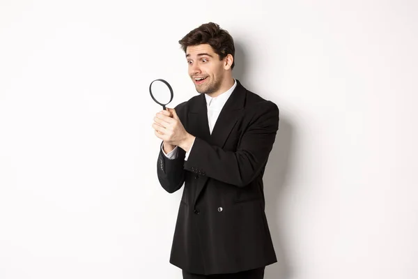 Image of handsome businessman in black suit, looking through magnifying glass, found something and smiling, standing over white background — Stock Photo, Image