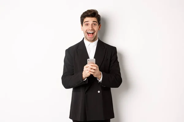Bild av glada stilig kille som får promo erbjudande på mobiltelefon, ser förvånad och ler, står mot vit bakgrund — Stockfoto