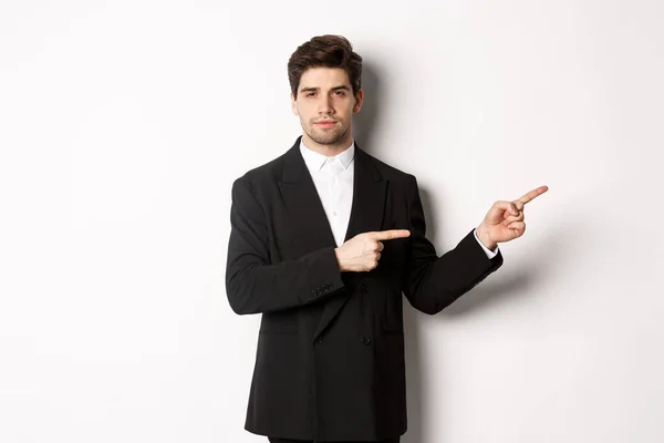 Bild av stilig affärsman i svart kostym, pekar finger höger och tittar på kameran, står mot vit bakgrund — Stockfoto