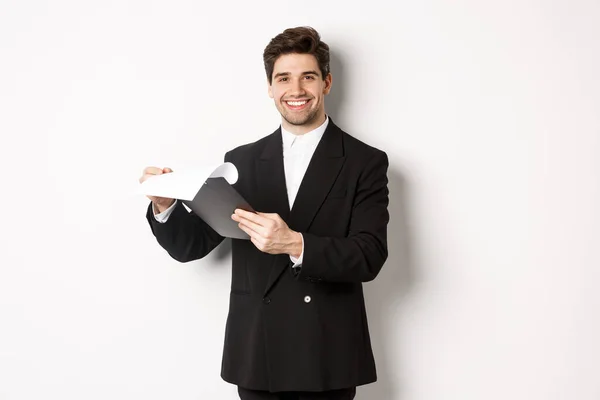 Beeld van een zelfverzekerde zakenman in pak die documenten bekijkt, klembord vasthoudt en blij lacht, tegen een witte achtergrond staat — Stockfoto