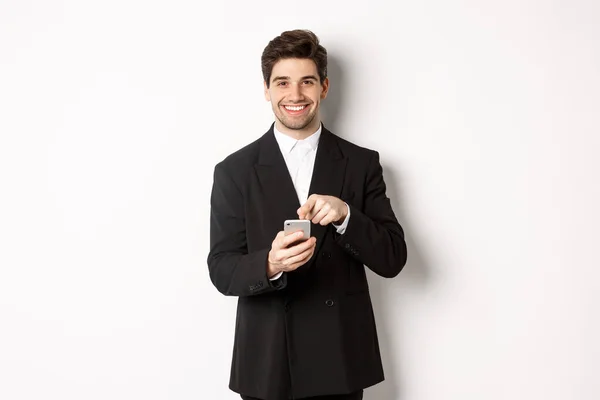 Wizerunek przystojnego biznesmena w garniturze pokazującego aplikację na smartfonie, wskazującego palcem na ekran komórkowy i uśmiechniętego, stojącego nad białym tłem — Zdjęcie stockowe