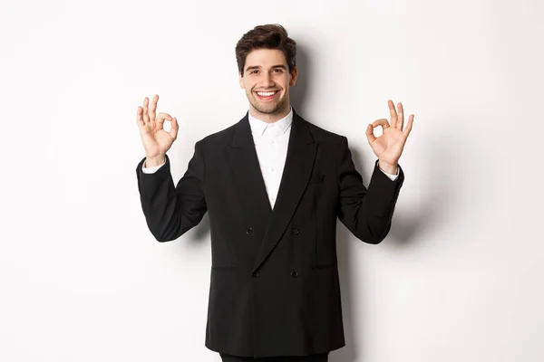 Concept of new year party, celebration and lifestyle. Portrait of handsome attractive man in black suit, smiling and showing okay signs, approve and recommend something, white background — Stock Photo, Image