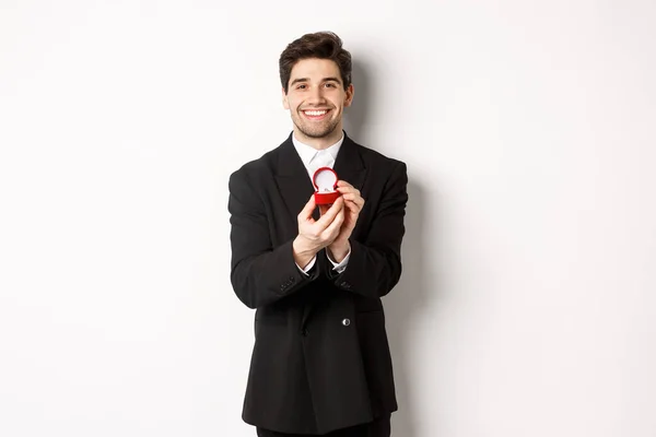 Image of handsome boyfriend in black suit making a proposal, asking to marry him and showing wedding ring, standing over white background — Stock Photo, Image