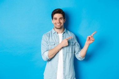 Çekici yetişkin bir adamın portresi gülümsüyor, parmaklarını logoya ya ya da afişe doğrultuyor, mavi arka plana karşı reklam gösteriyor.