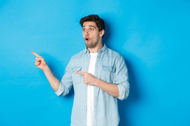 Şaşırmış ve şaşırmış bir adam terfiye bakıyor, parmaklarını reklamlara gösteriyor, mavi arka planda duruyor.