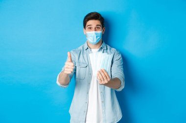 Koronavirüs, karantina ve sosyal uzaklık kavramı. Tıbbi maskeler ve başparmak gösteren genç adam, mavi arka planda duruyor.