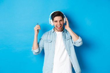 Mutlu gülümseyen adam kulaklıkla müzik dinlemekten zevk alıyor, akıllı telefonu havada tutuyor, mavi arka planda duruyor.