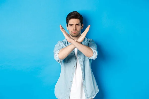 Displeased man frowning and saying no, making cross sign, tell to stop, standing against blue background