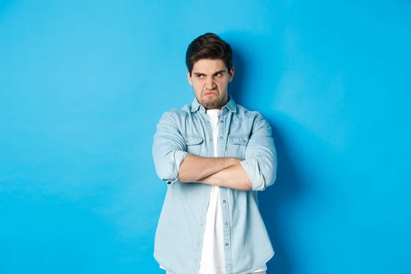 Angry guy cross arms on chest and looking away with insulted expression, standing offended against blue background
